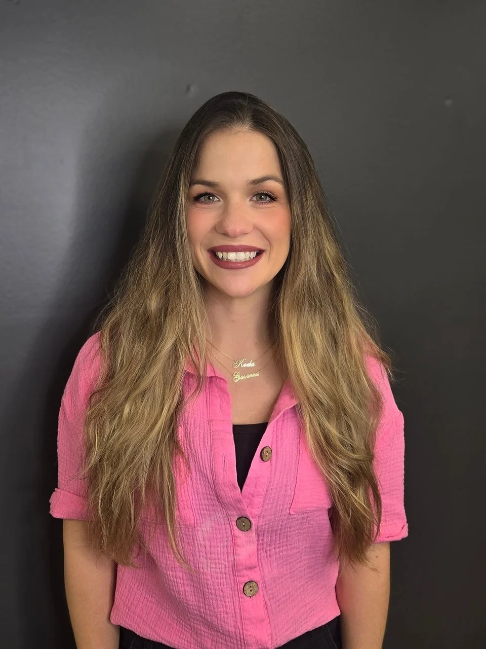 A young woman with long wavy hair and braces, smiling, wearing a pink button-up shirt with rolled-up sleeves, a black top underneath, and a gold necklace with a name pendant, standing against a dark gray background.