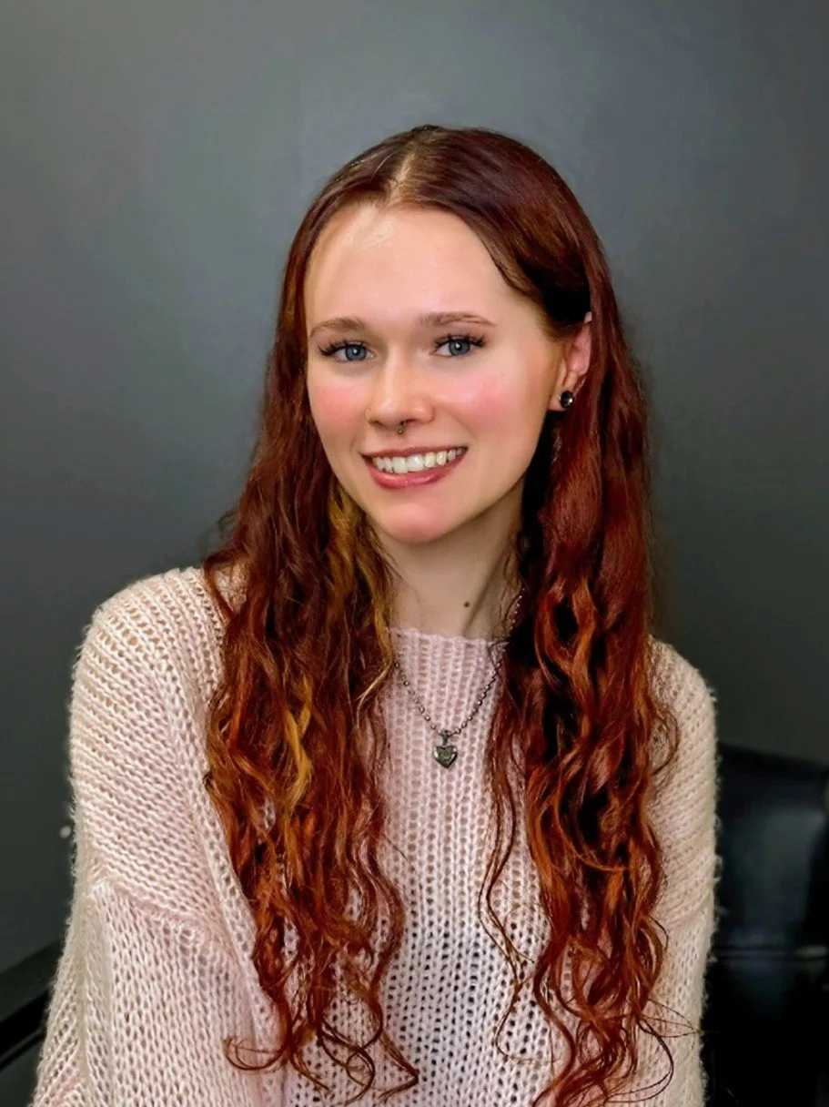 Portrait of a young woman with long, curly red hair, wearing a pink knitted sweater, black earrings, and a necklace with a pendant, smiling at the camera against a plain dark grey background.
