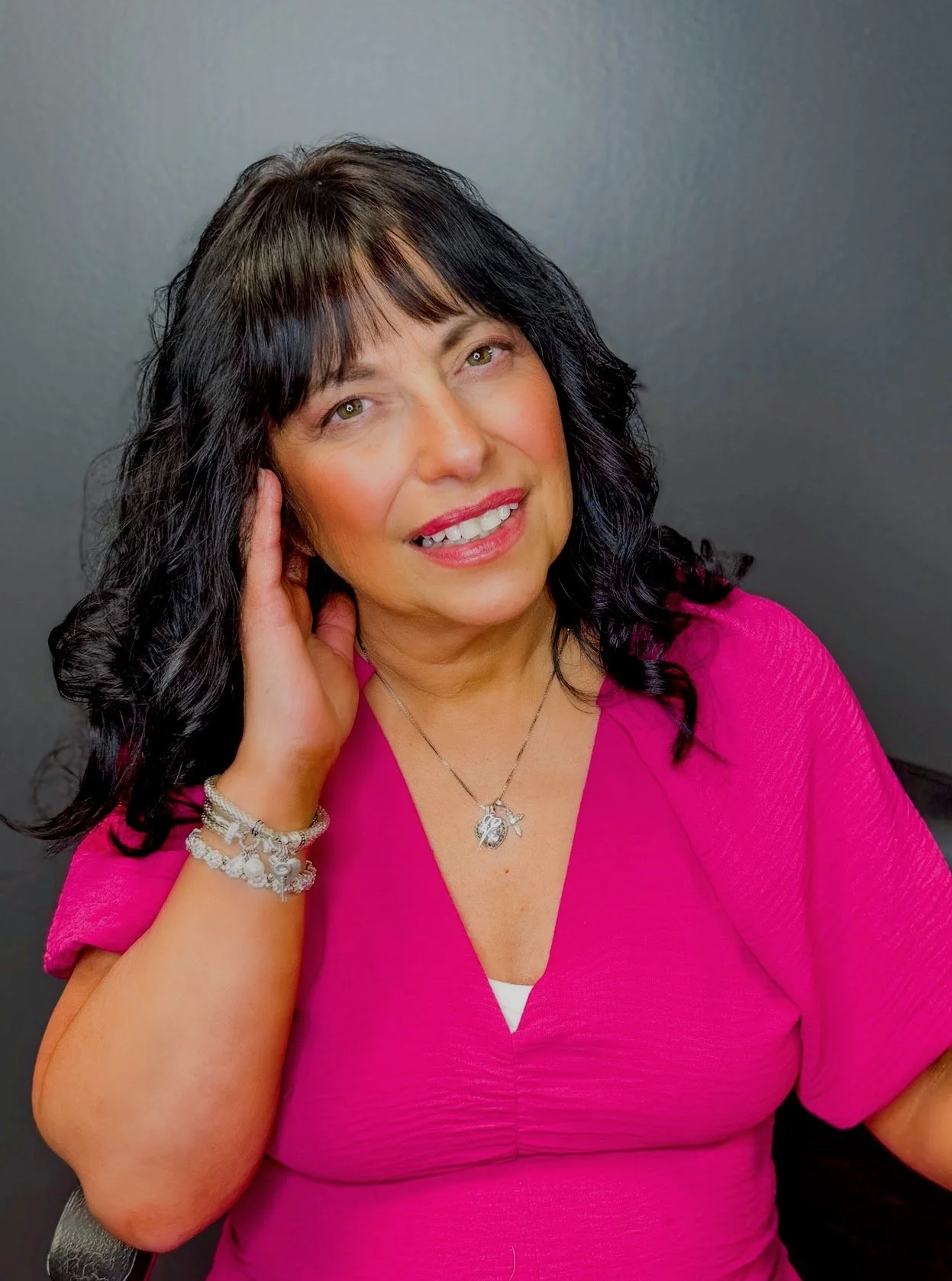 A woman with black hair, wearing a pink top, smiling with her hand touching her face, against a gray background.