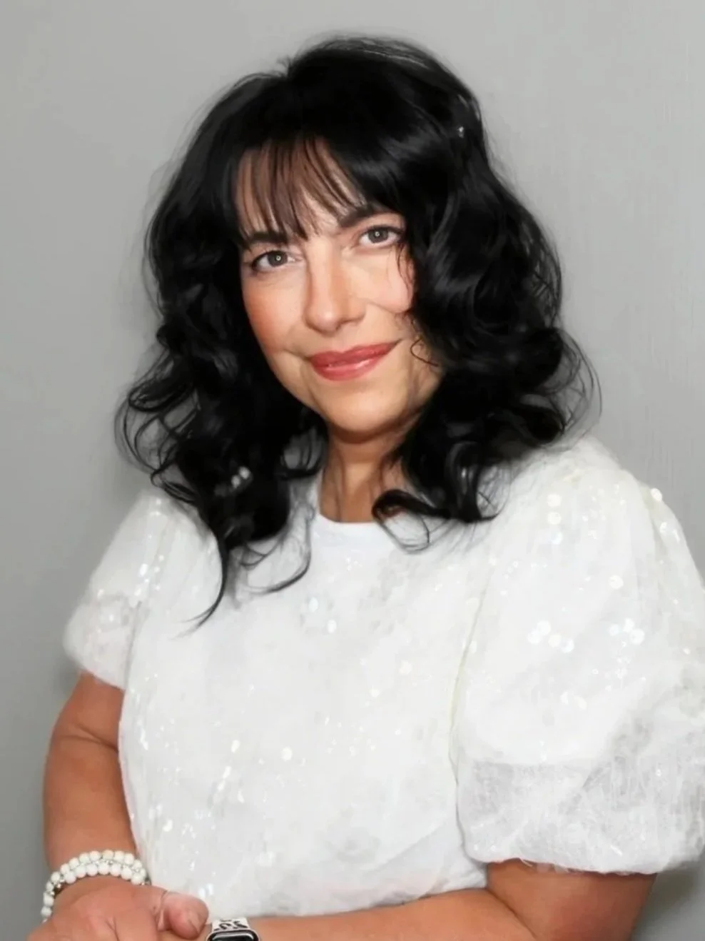 A woman with black hair, wearing a pink top, smiling with her hand touching her face, against a gray background.