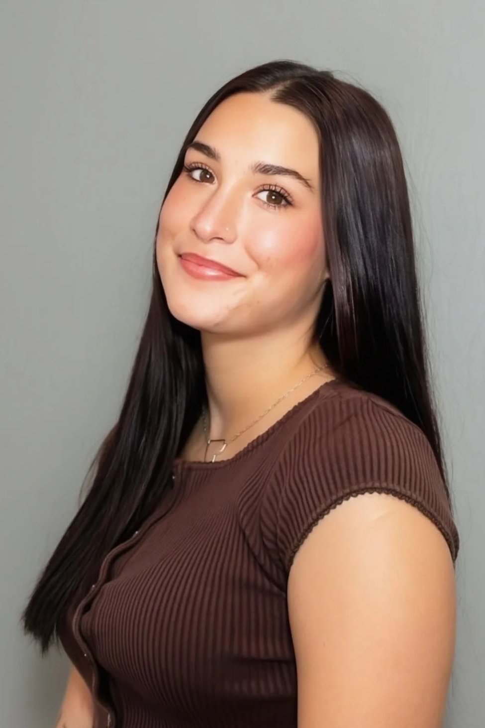 Close-up of a woman with long black hair, fair skin, wearing makeup, a nose piercing, smiling slightly, wearing a black top and a light-colored patterned jacket against a gray background.