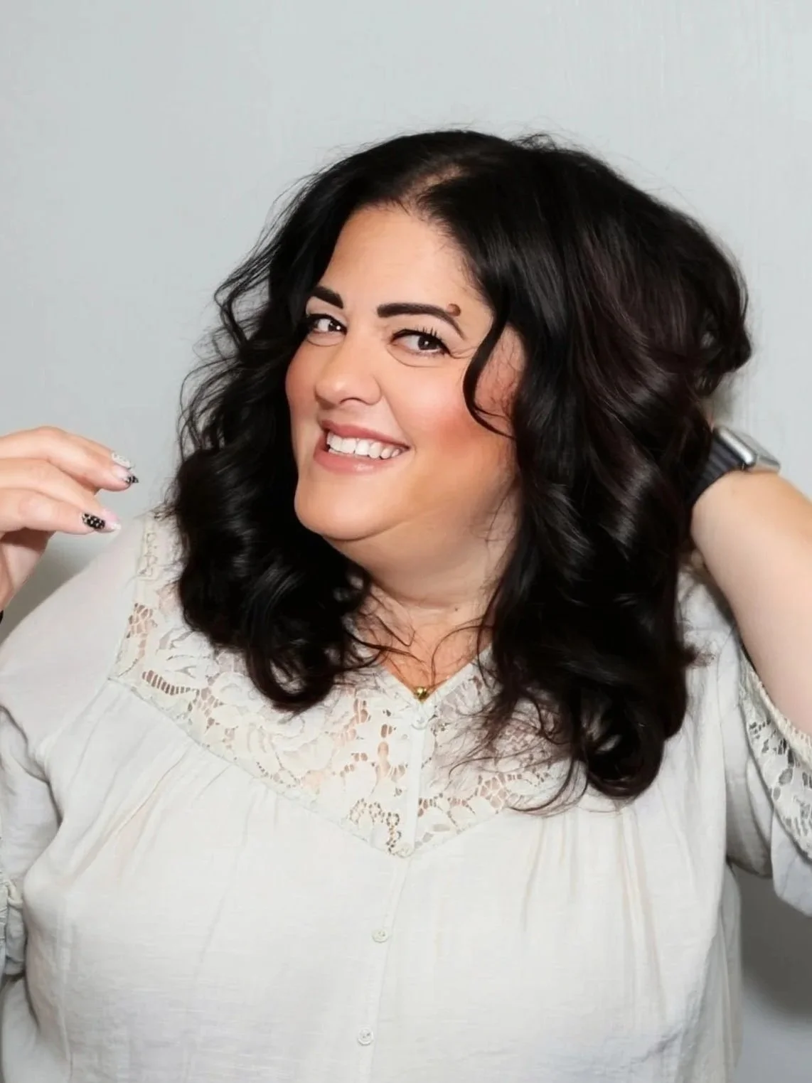 A woman with shoulder-length curly dark hair, wearing a pink top and gold jewelry, smiling against a dark gray background