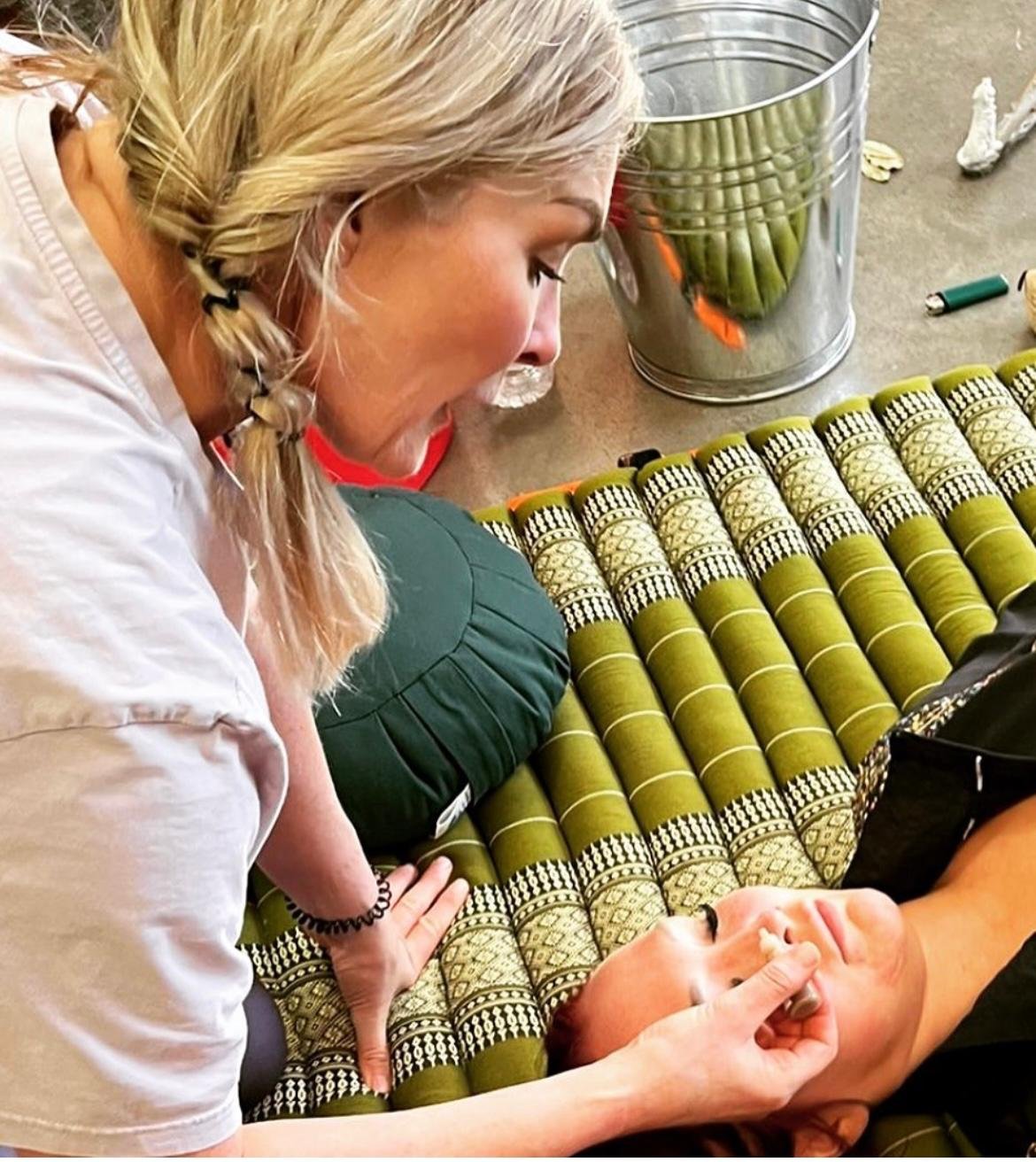 Person lying on a patterned mat receiving a facial treatment, another person crouched beside them performing the procedure.
