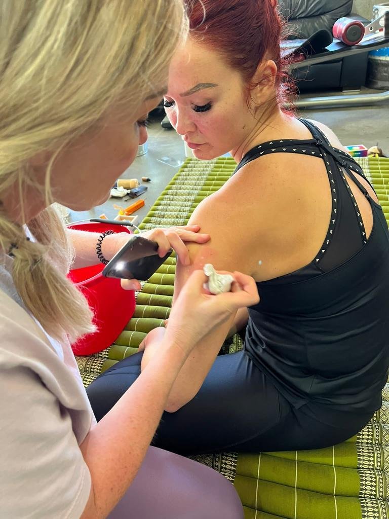 Two women during a tattooing or skin art session, one applying design to the other's shoulder with a tool.