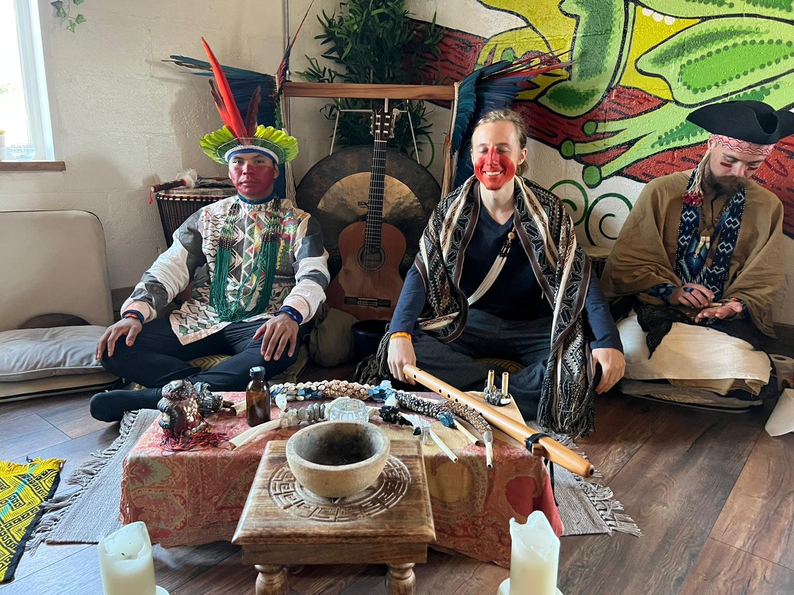 Three people sitting indoors in a ceremonial setting with musical instruments, candles, and vibrant decorations; each wearing traditional or indigenous-style clothing and face paint.