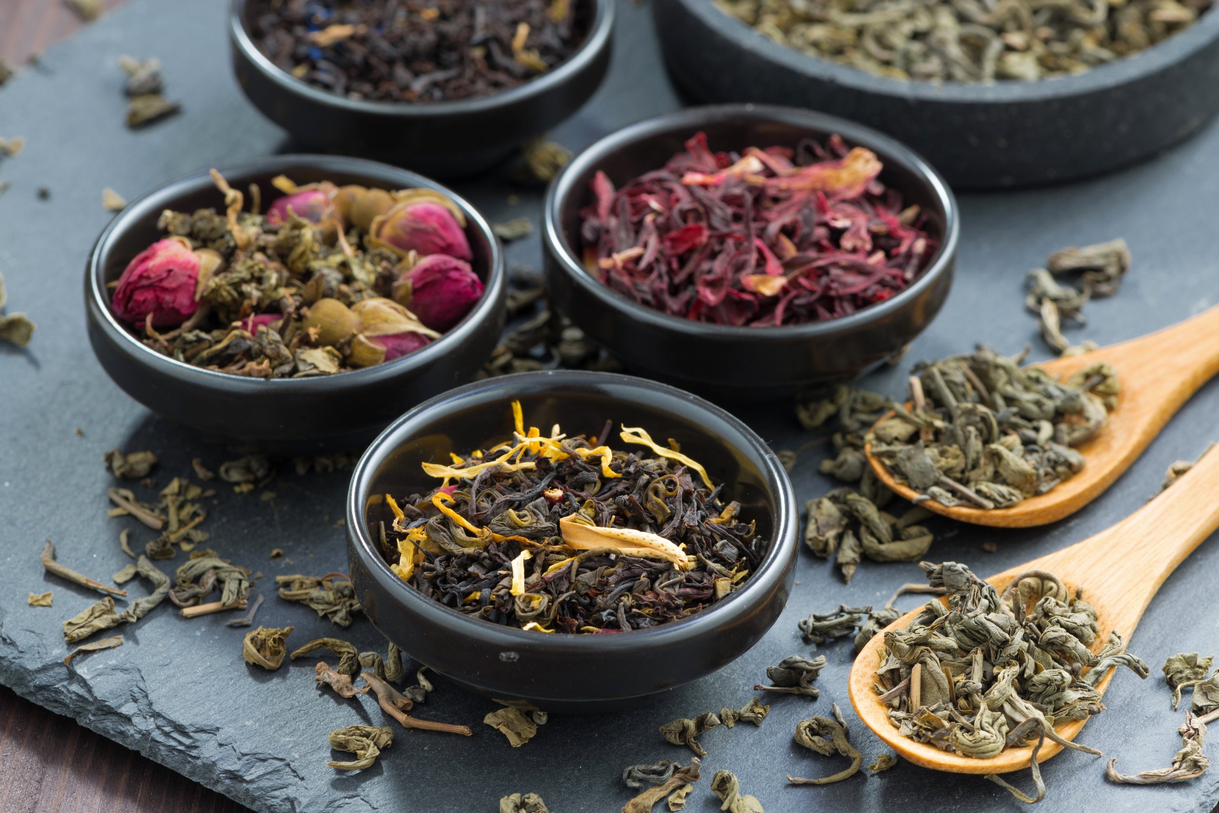 Assorted dried loose leaf teas and herbs in black bowls on a slate surface, with tea leaves on wooden spoons.