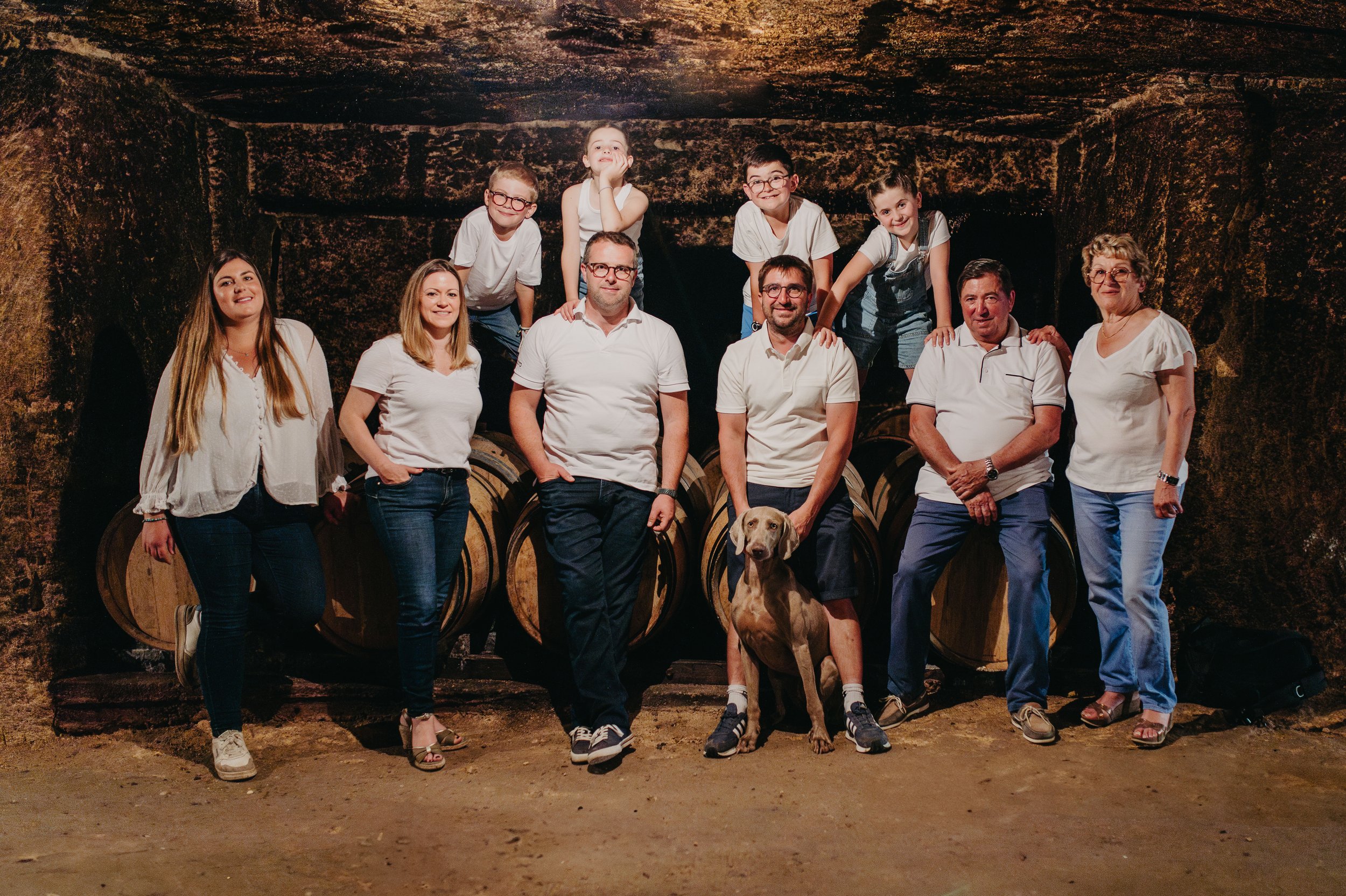 Un groupe de personnes posant devant des tonneaux dans une cave, avec un chien assis au premier plan.