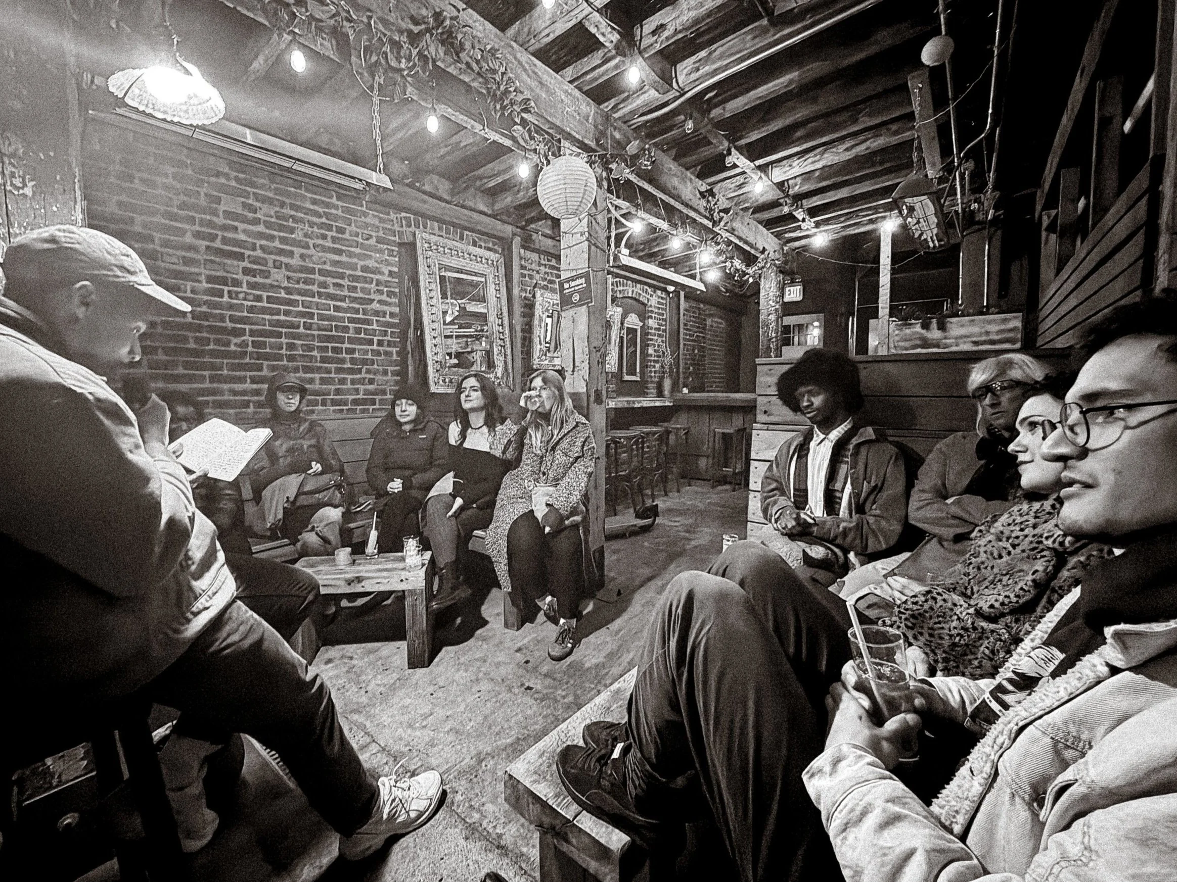 A group of people sitting on benches and chairs around a person reading from a book in a cozy, rustic cafe with brick walls, hanging paper lanterns, and wood decor.