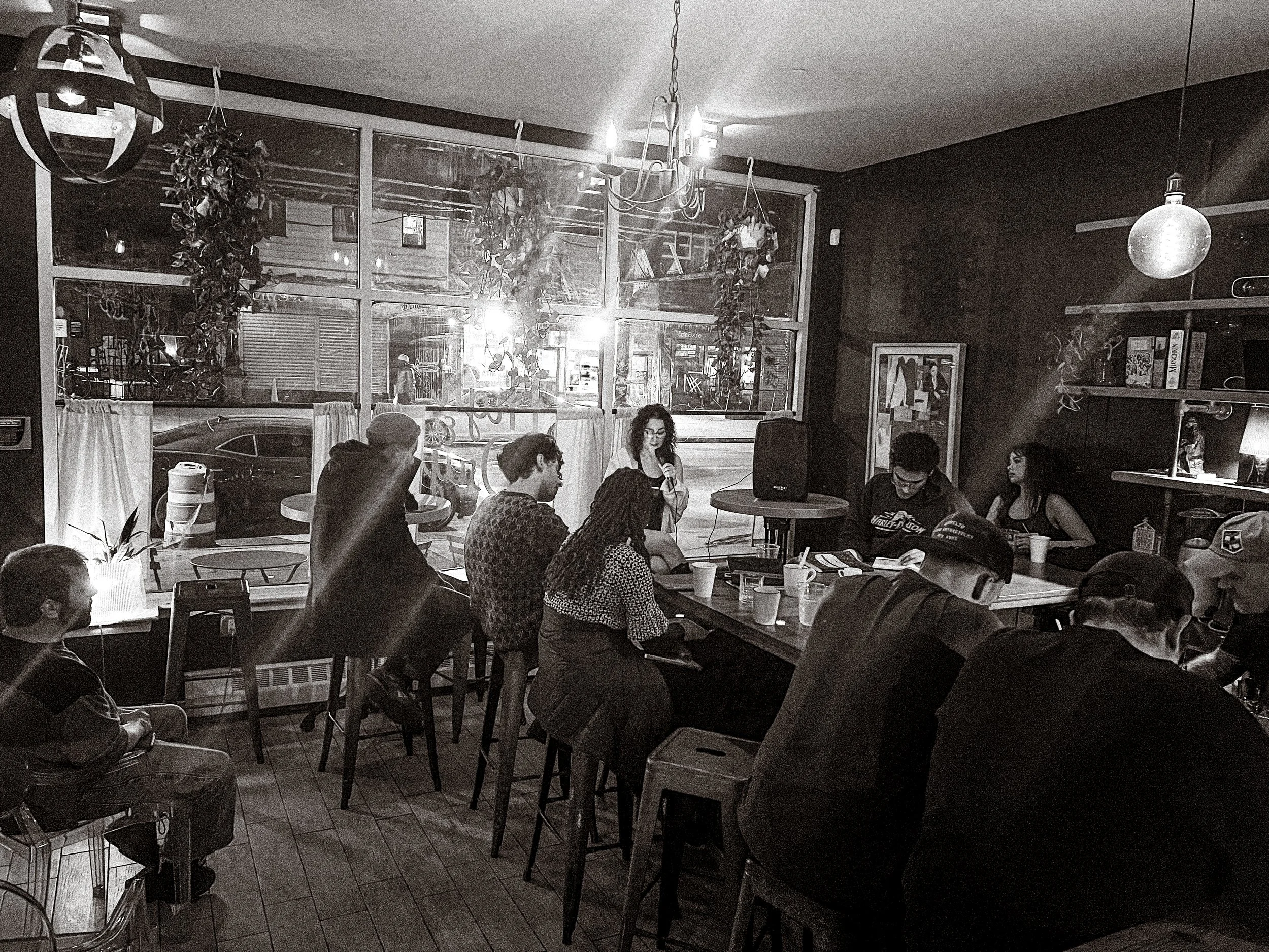 People gathered in a cozy cafe, sitting at tables and listening to a woman speaking into a microphone near a window with street view at night. The cafe is decorated with hanging plants, bookshelves, and warm lighting, creating an intimate atmosphere.