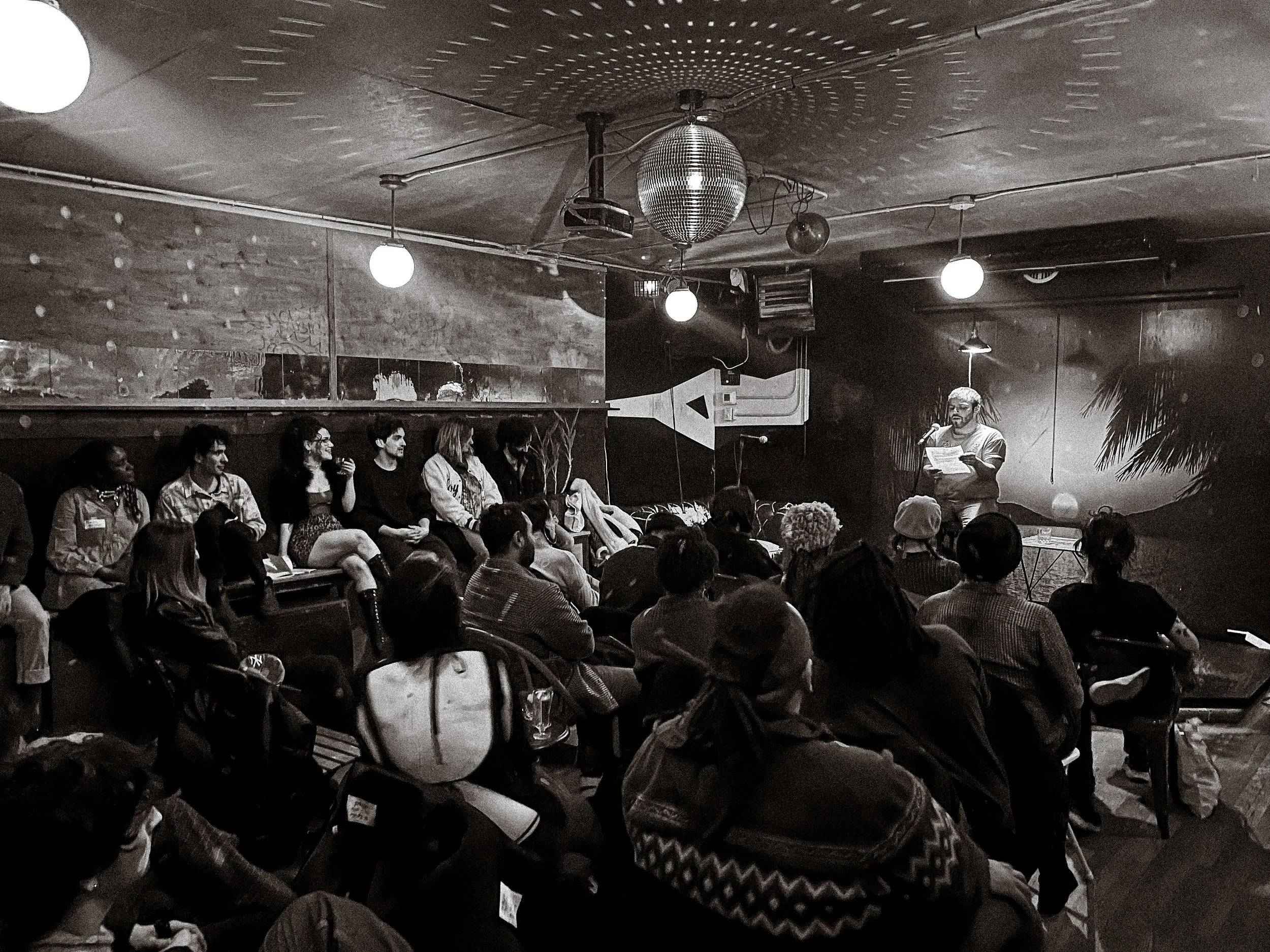 A black and white photo of a person reading from a paper on stage in front of an audience in a dimly lit room decorated with a disco ball and hanging spherical lights.