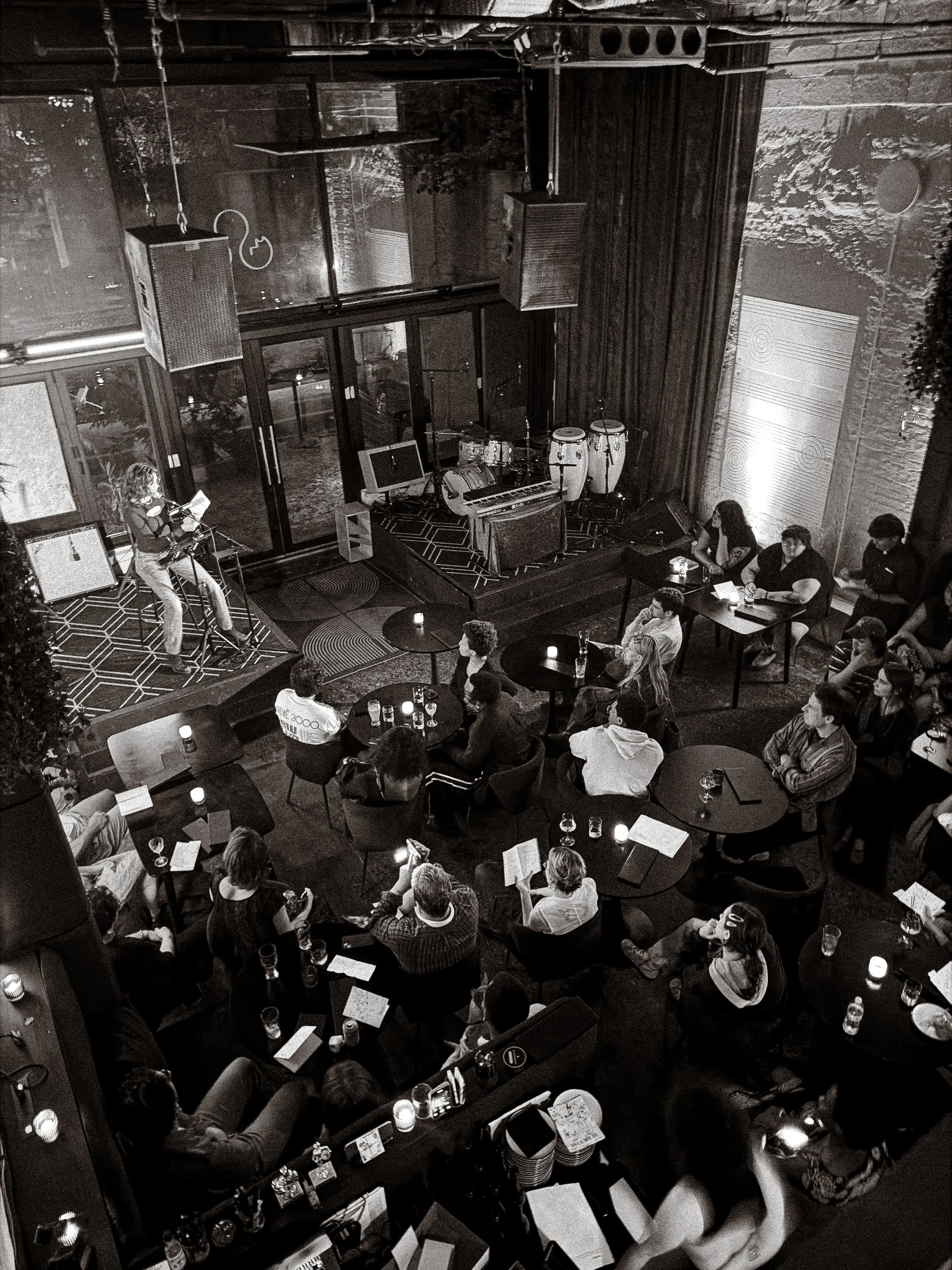 A black-and-white photo a poetry reading where an audience of diverse people is seated at tables, watching and listening. 