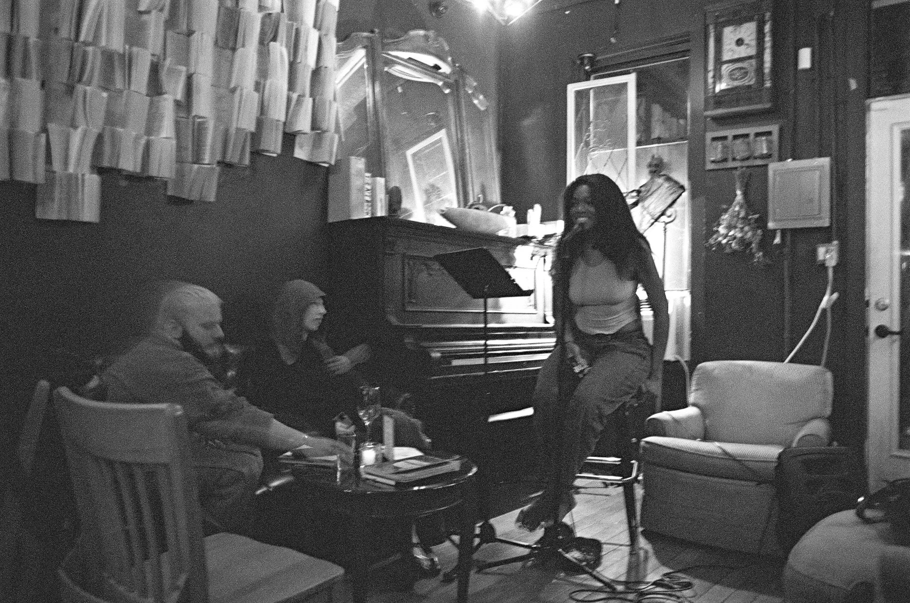 A woman reading in front of an audience in a cozy, dimly lit room with musical instruments, books, and artwork.