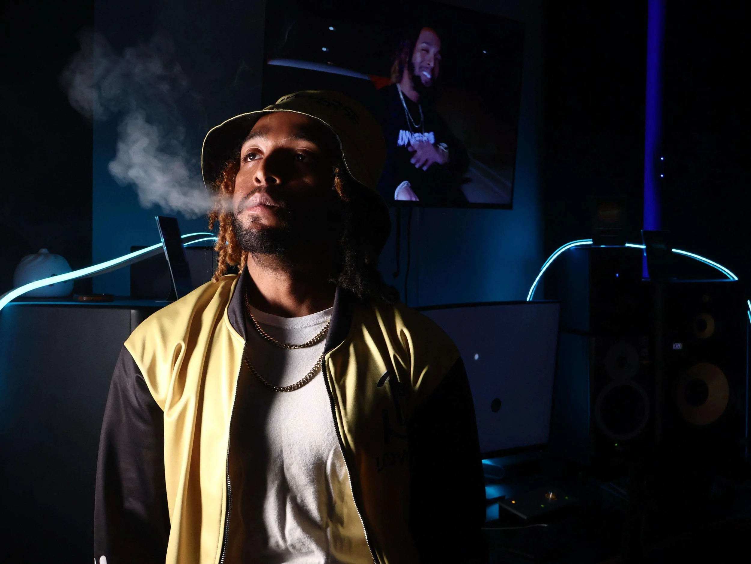 A man with dreadlocks and a beard wearing a tan bucket hat, white t-shirt, and yellow jacket, smoking and looking serious in a dark room with blue neon lighting. There is a large screen in the background displaying his face and a DJ setup with turntables.