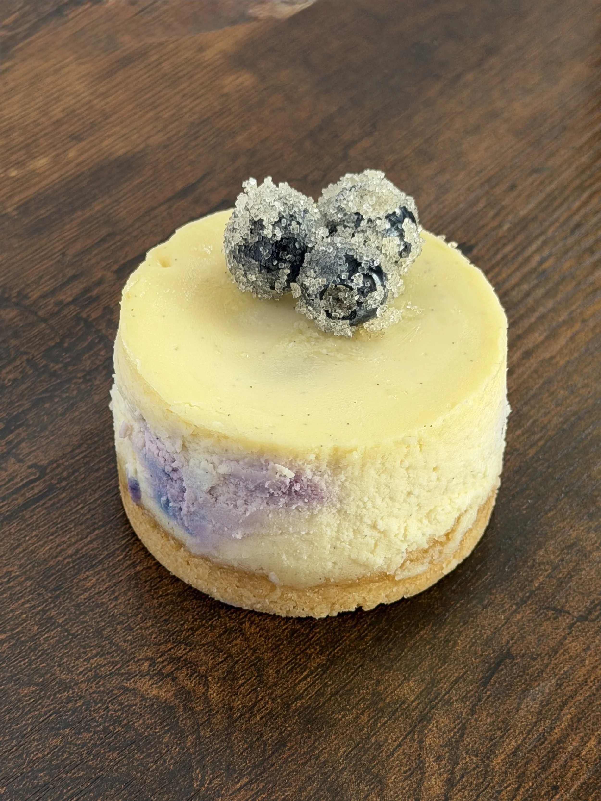 Slow-baked Tall Blueberry New York-Style Vanilla Bean cheesecake crafted with premium cream cheese, local farm eggs, and real vanilla bean. Features whole blueberries baked throughout, a buttery vanilla cookie crust, and seasonal sugared local bluebe