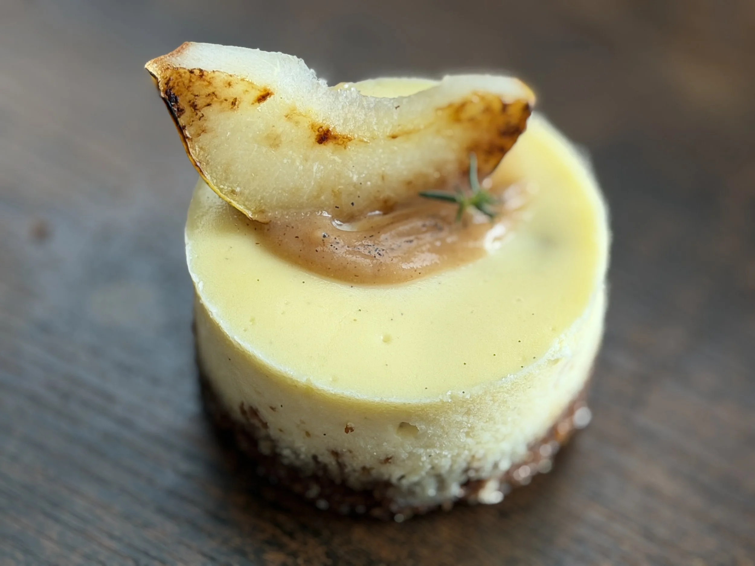 Tall New York-Style Vanilla Bean Cheesecake with a homemade gingersnap cookie crust. Topped with sautéed pear and delicate pear coulis, then finished with a light sprinkle of vanilla bean and an aromatic herb sprig.
