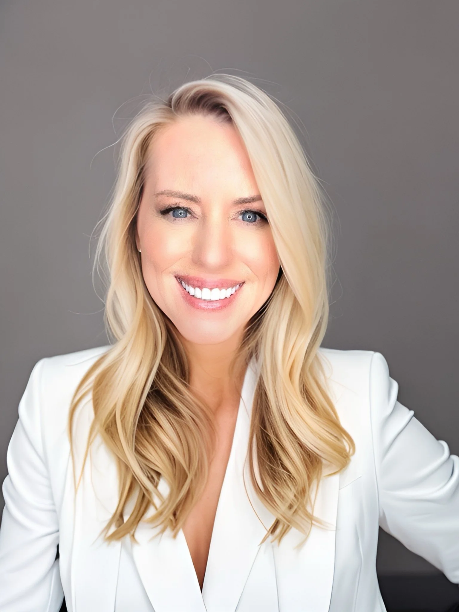 A smiling woman with long blonde hair, wearing a white blazer, against a gray background.