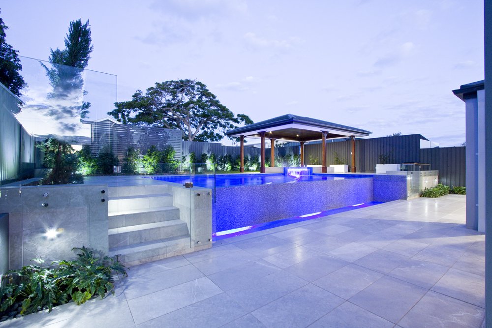 Modern backyard with a swimming pool, outdoor seating area with wooden posts, and a covered pavilion at dusk.