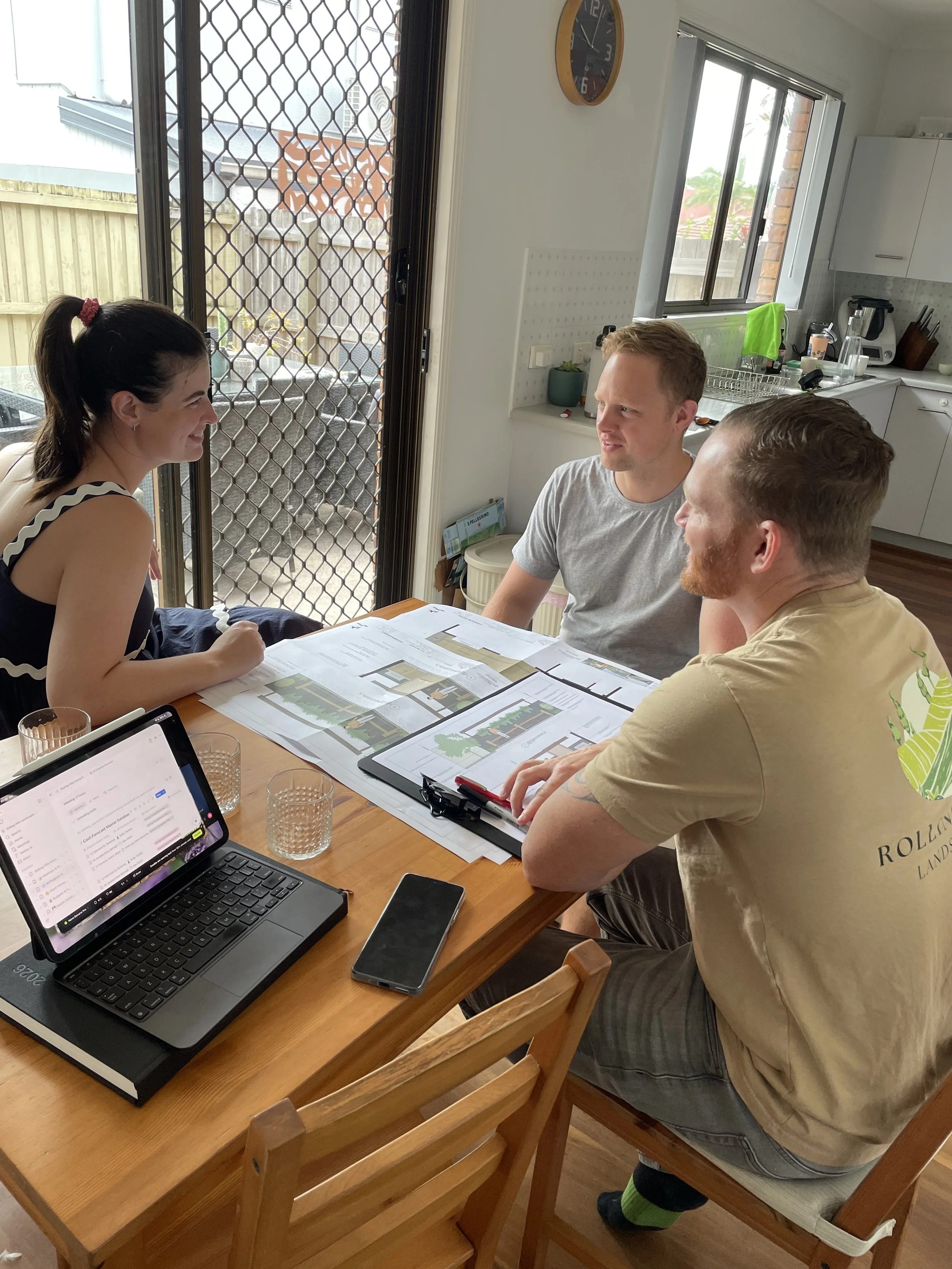 Three people sitting at a dining table, discussing architectural plans, with a laptop and a tablet, in a kitchen with a sliding door to an outdoor area.