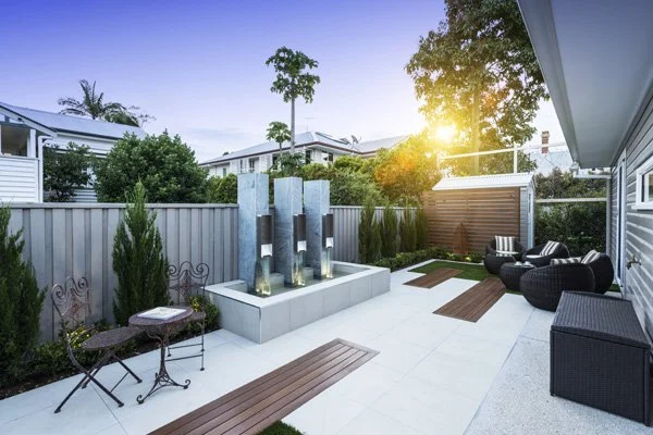 Modern backyard patio area with outdoor seating, water feature, and lush greenery at sunset.