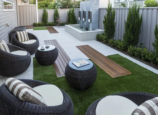 Modern backyard patio with black wicker chairs, matching tables, striped cushions, wooden pathway, and a water feature with a fountain, surrounded by green plants and trees.