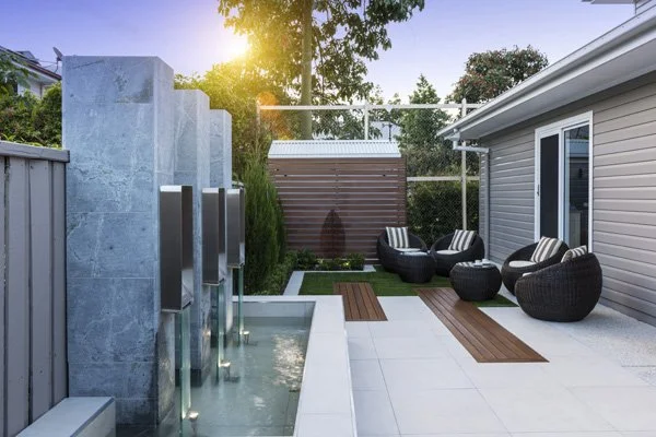 Modern backyard patio with a water feature, outdoor seating with rattan chairs and tables, and a wooden pathway, surrounded by a fence, house, and trees.