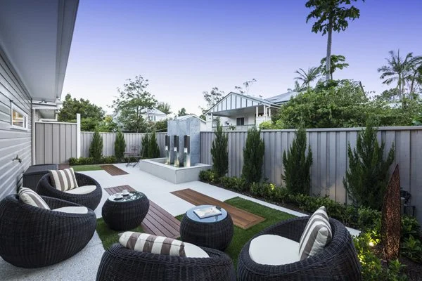 Backyard patio with black wicker outdoor furniture with striped cushions, a fountain feature with three water spouts, a garden with tall shrubs, and a gray fence surrounding the yard.
