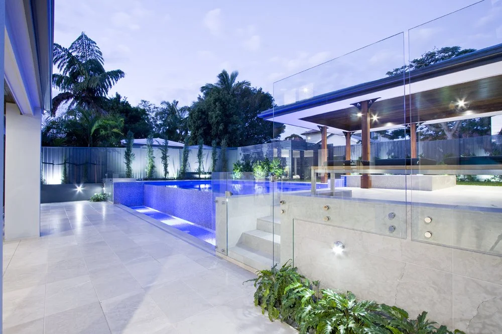 Modern outdoor backyard with a lit swimming pool, glass fence, and a covered patio area with wooden columns, surrounded by trees and plants.