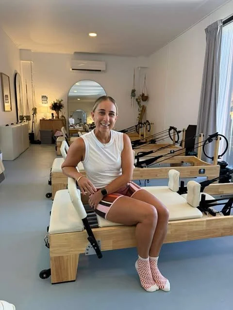 A pilates instructor sitting on a Pilates reformer machine in a bright, modern studio with other reformers and decor around.