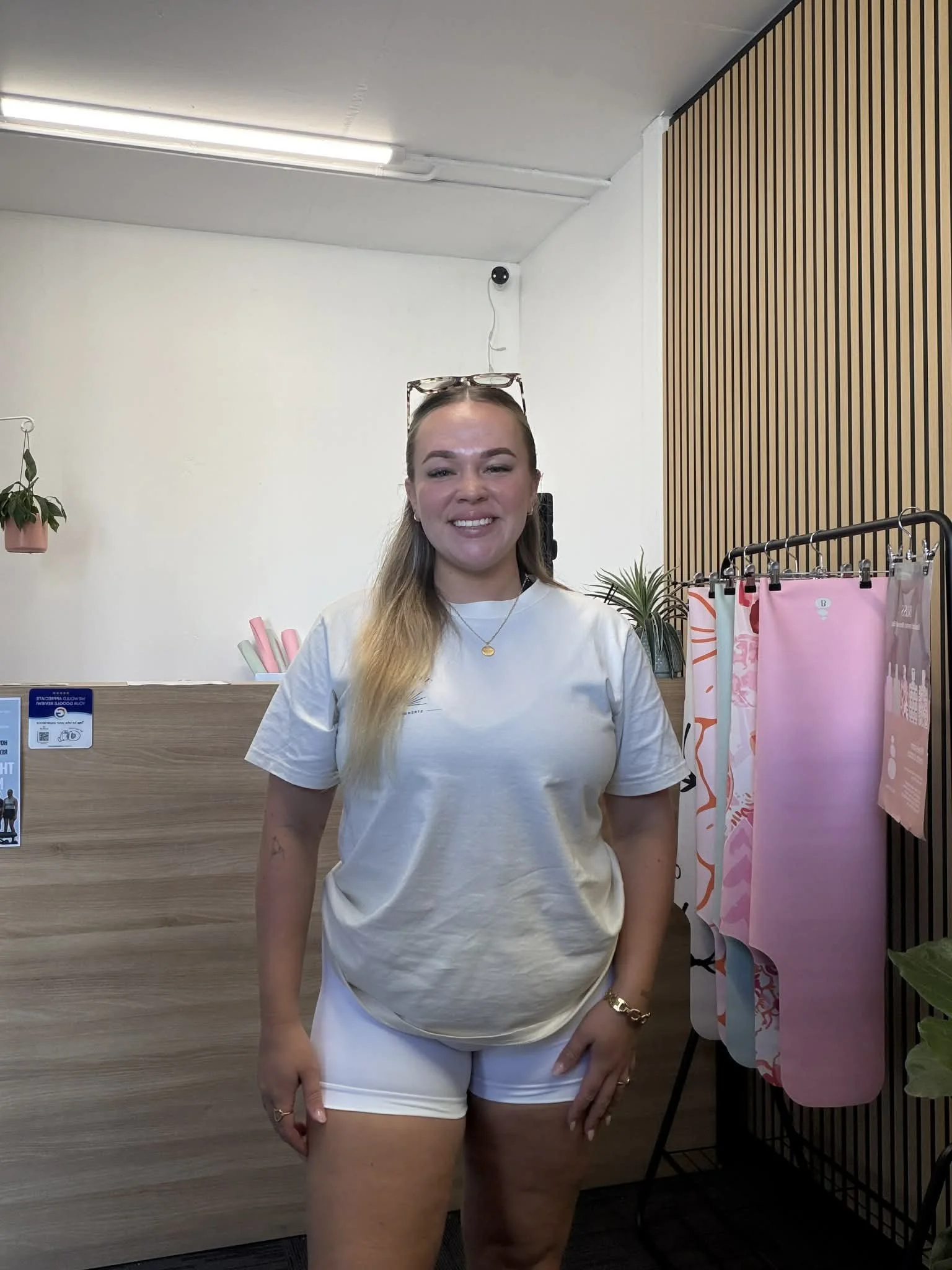 Smiling gym receptionist, standing indoors near a wooden counter and a rack of pink and patterned pilates mats and socks.