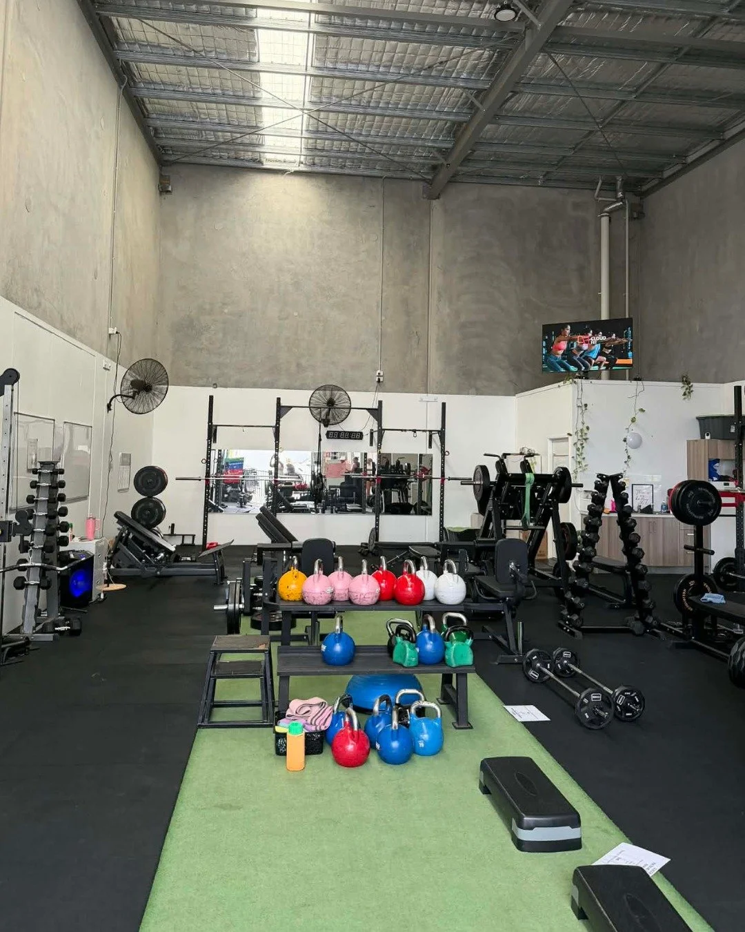 A well-equipped gym with kettlebells, weights, benches, and exercise machines, with a large mirror, fans, a television, and a green turf area for workouts.