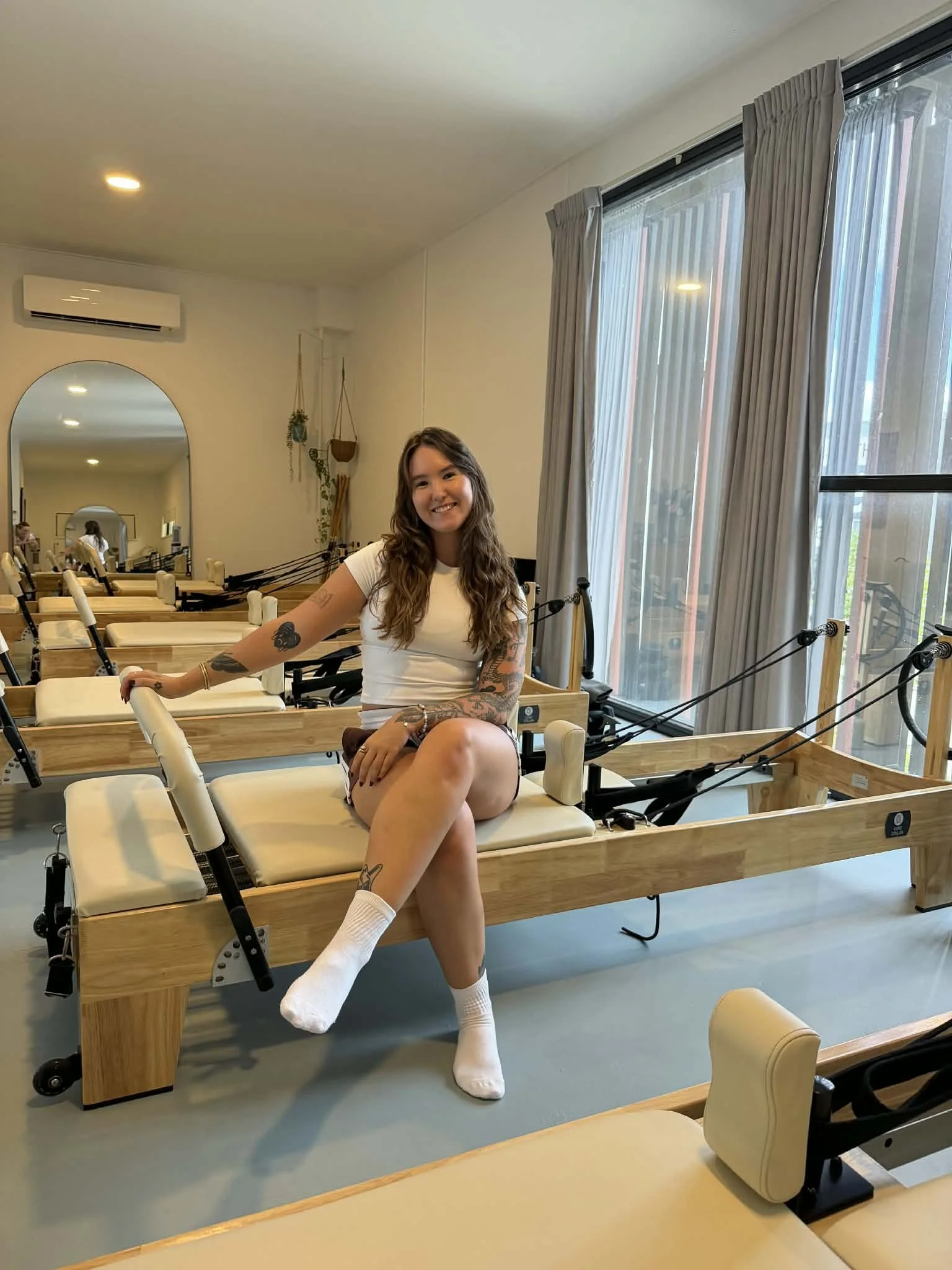 A pilates instructor sitting on a Pilates reformer machine in a bright, modern studio, smiling at the camera.