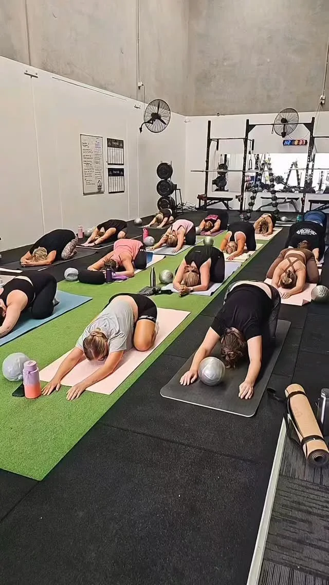 A group of women participating in a mat pilates class, practicing Child's Pose on mats in a gym or fitness studio.