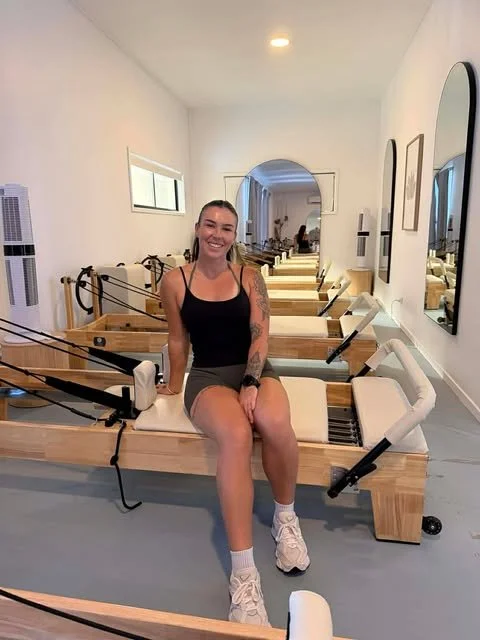 A pilates instructor sitting on a Pilates reformer machine in a ballet studio with mirrors and windows.