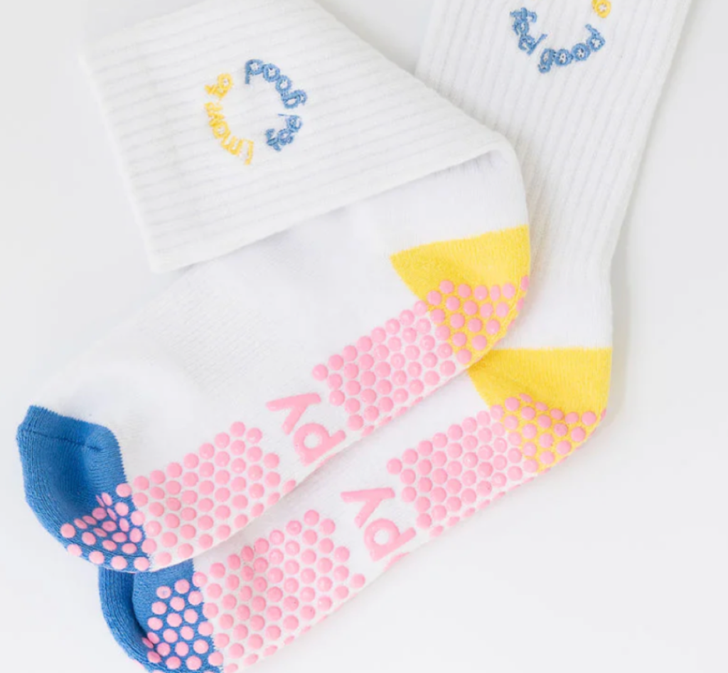 Pair of white socks with non-slip pink grips, blue toe, yellow heel, and embroidered text 'feel good' and 'yoga' on the top.