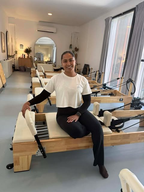 A pilates instructor smiling and sitting on a Pilates reformer machine in a fitness studio.