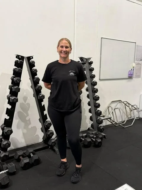 A gym instructor standing in gym, surrounded by workout equipment.