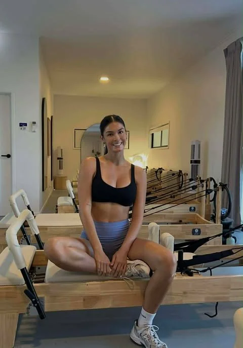 A pilates instructor sitting on a Pilates reformer machine in a fitness studio, smiling at the camera, wearing a black sports bra, gray shorts, and sneakers.