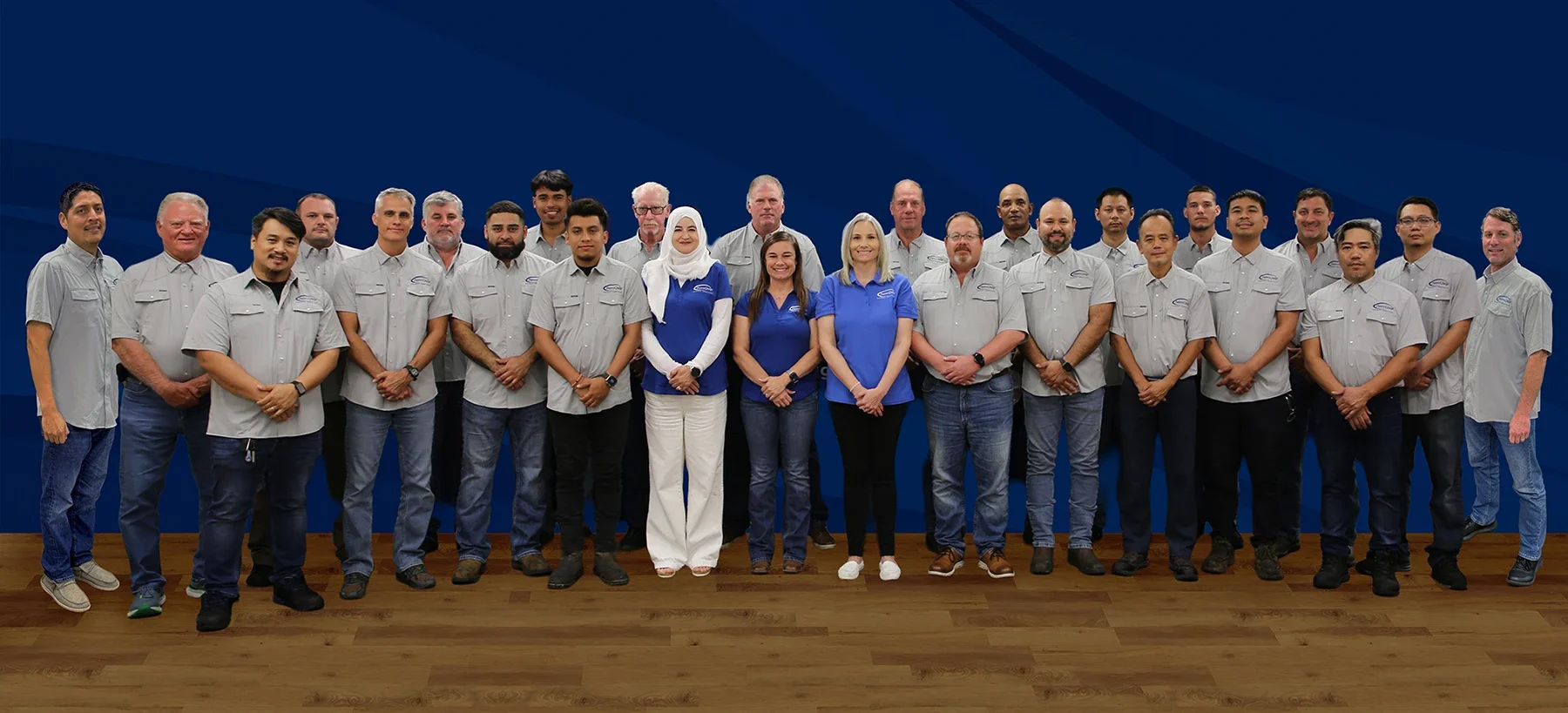 Group photo of diverse men and women wearing matching shirts, standing on a wooden floor against a blue background.