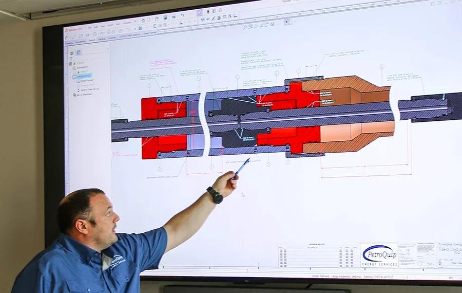A PetroQuip engineer in a blue company shirt is pointing at a technical engineering diagram of a tool component displayed on a large screen during an engineering design meeting.