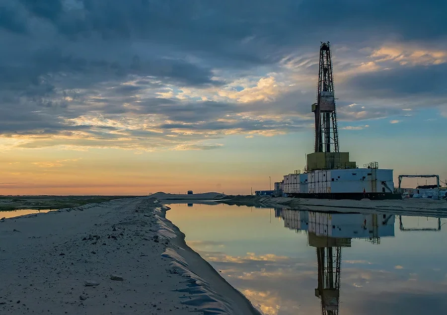 An oil drilling rig next to a waterway at sunset with a partly cloudy sky and reflected clouds in the water.