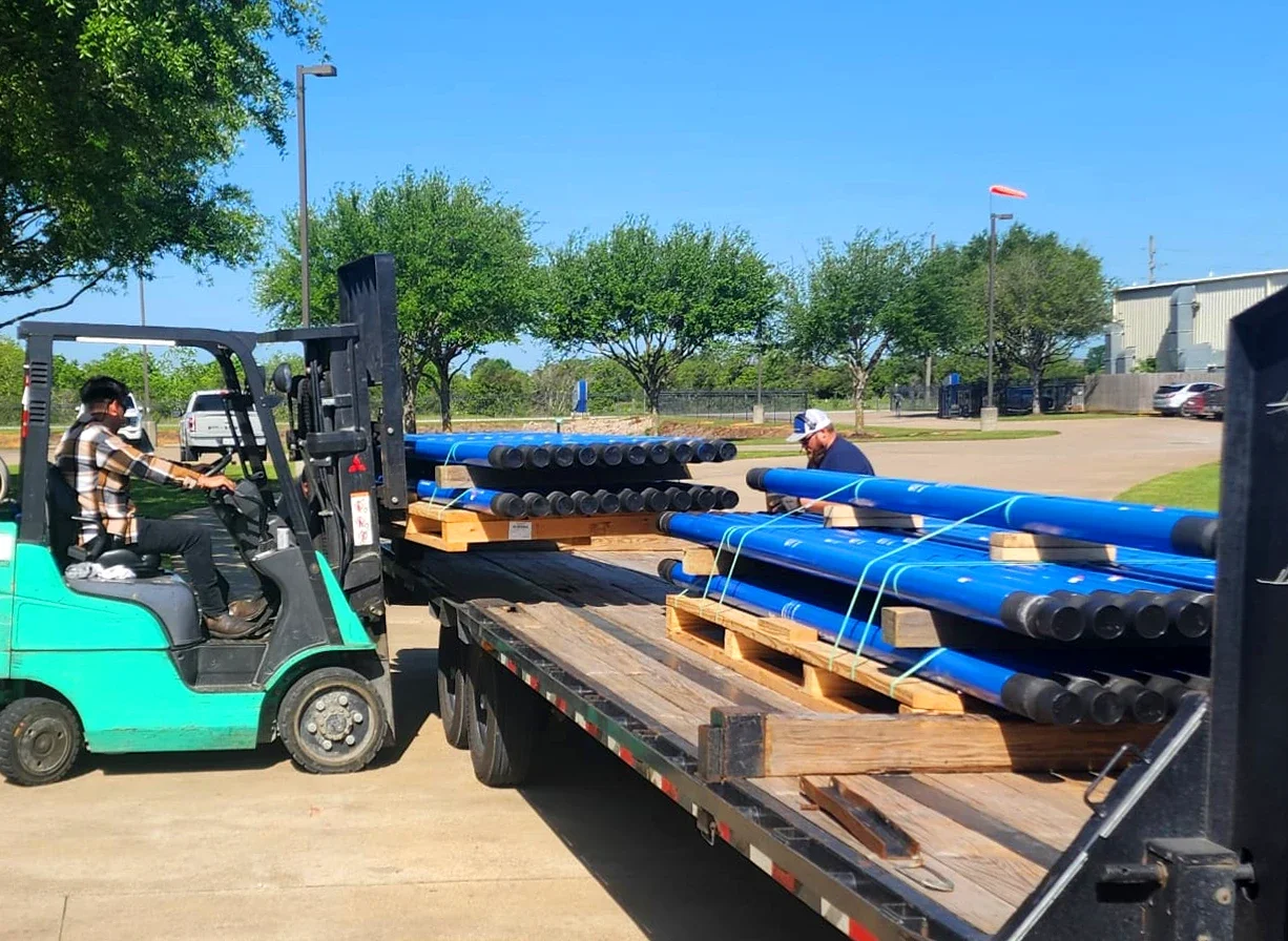 A person operating a forklift to unload or load long blue pipes onto a truck with a second person handling the pipes on the truck. The background shows trees, a parking lot, and a clear blue sky.