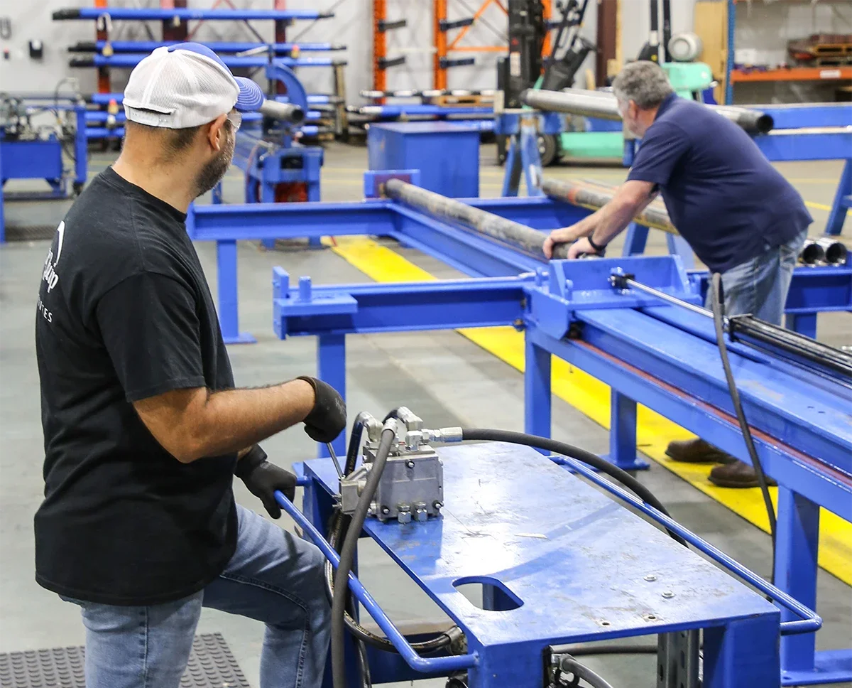 Two workers in a factory assembling or working with metal pipes and blue machinery in a workshop.