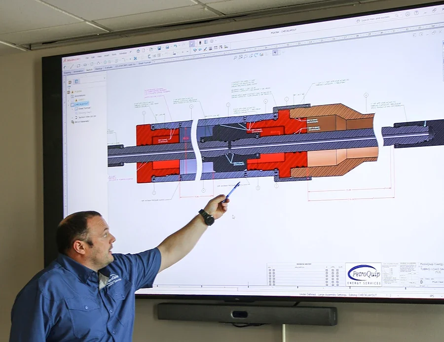 Man in blue shirt pointing at technical engineering drawing on a large screen, detailing a cross-section of a mechanical component or assembly.