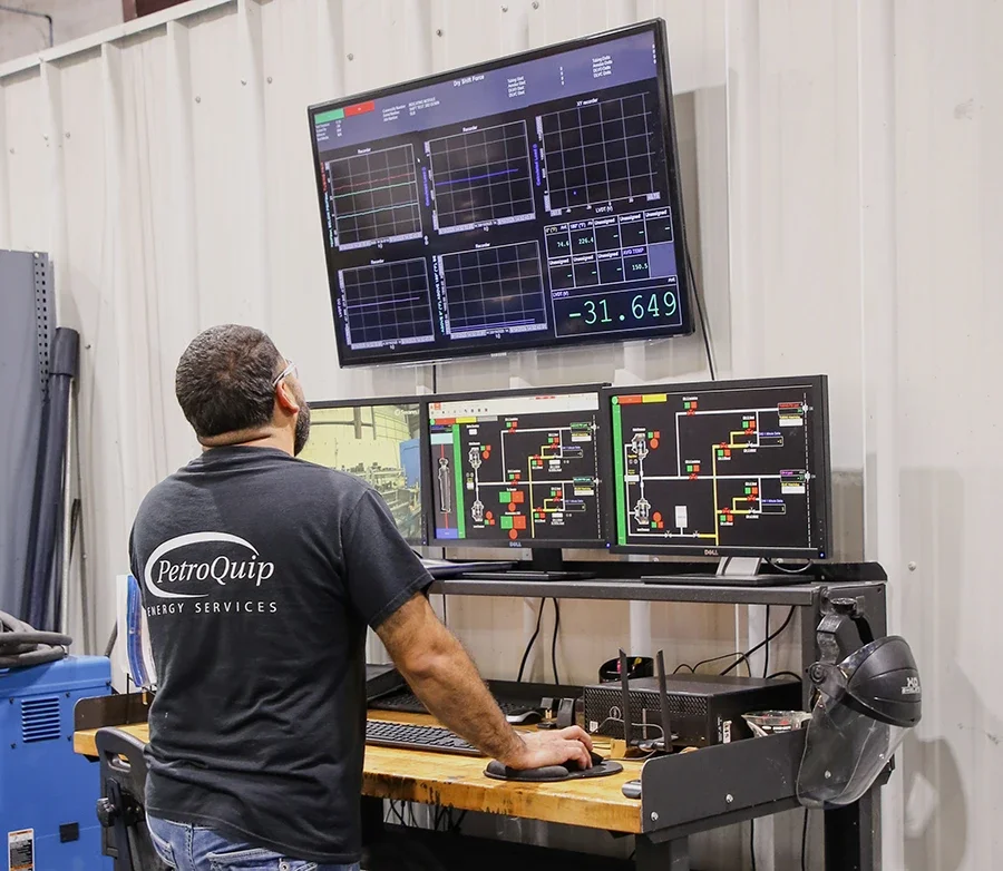 A PetroQuip team member working at a computer station with three monitors displaying data and process diagrams, wearing a black T-shirt with 'PetroQuip Energy Services' on the back, in a testing facility.