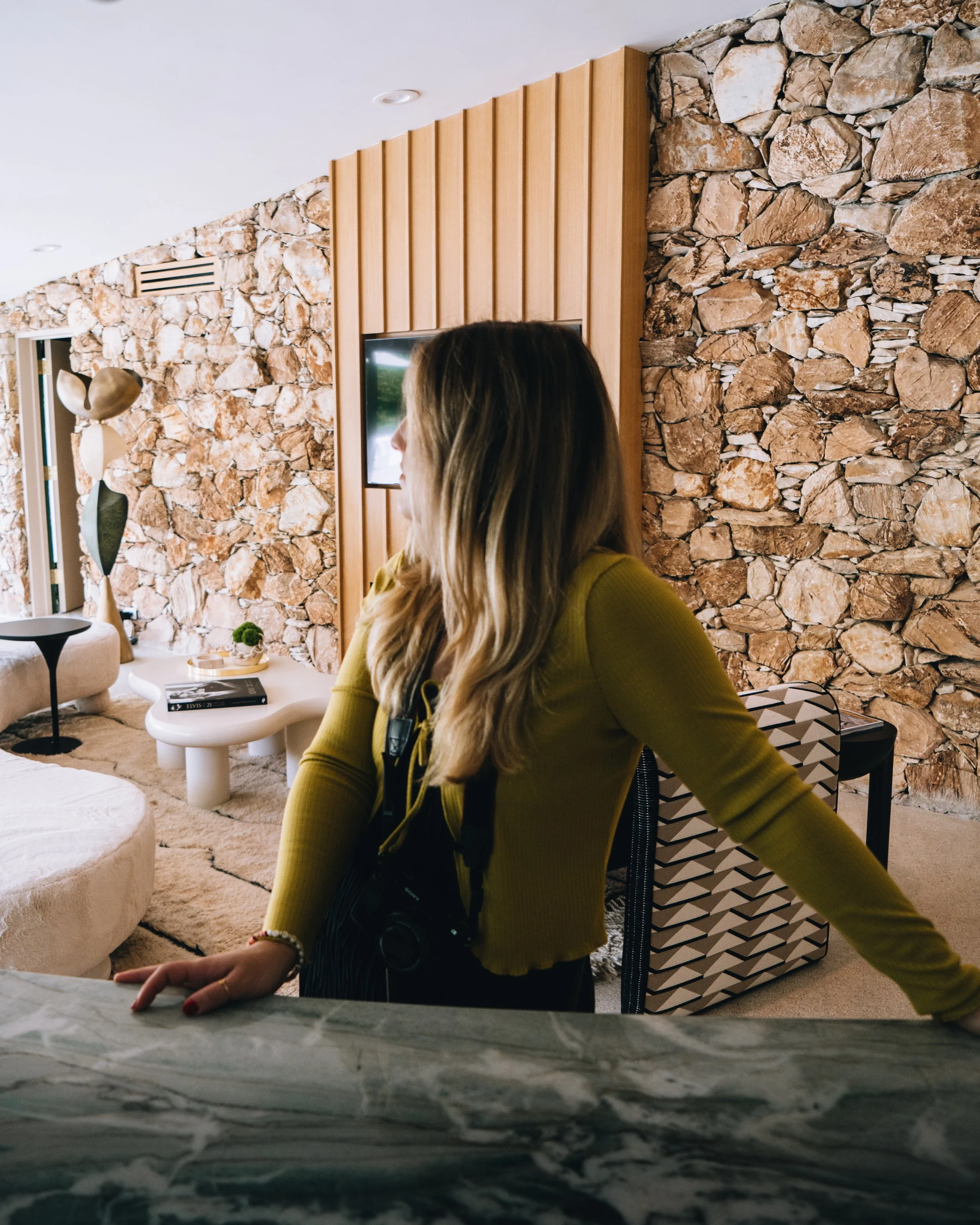 A woman with long blonde hair wearing a yellow top, leaning on a kitchen counter, with a camera hanging from her neck, in a modern living room with stone and wood wall decor of a Mid Century Modern home in Palm Springs.