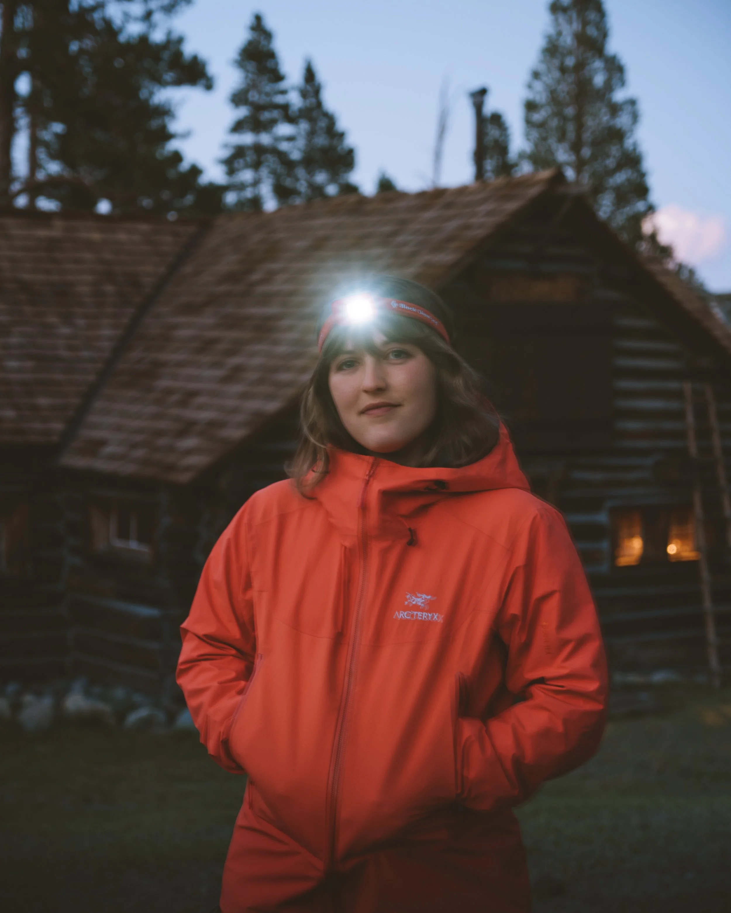 A young woman in a red Arc'teryx jacket and headlamp stands outdoors in front of a rustic cabin at dusk in Mammoth Mountain. Exploring in the dark.