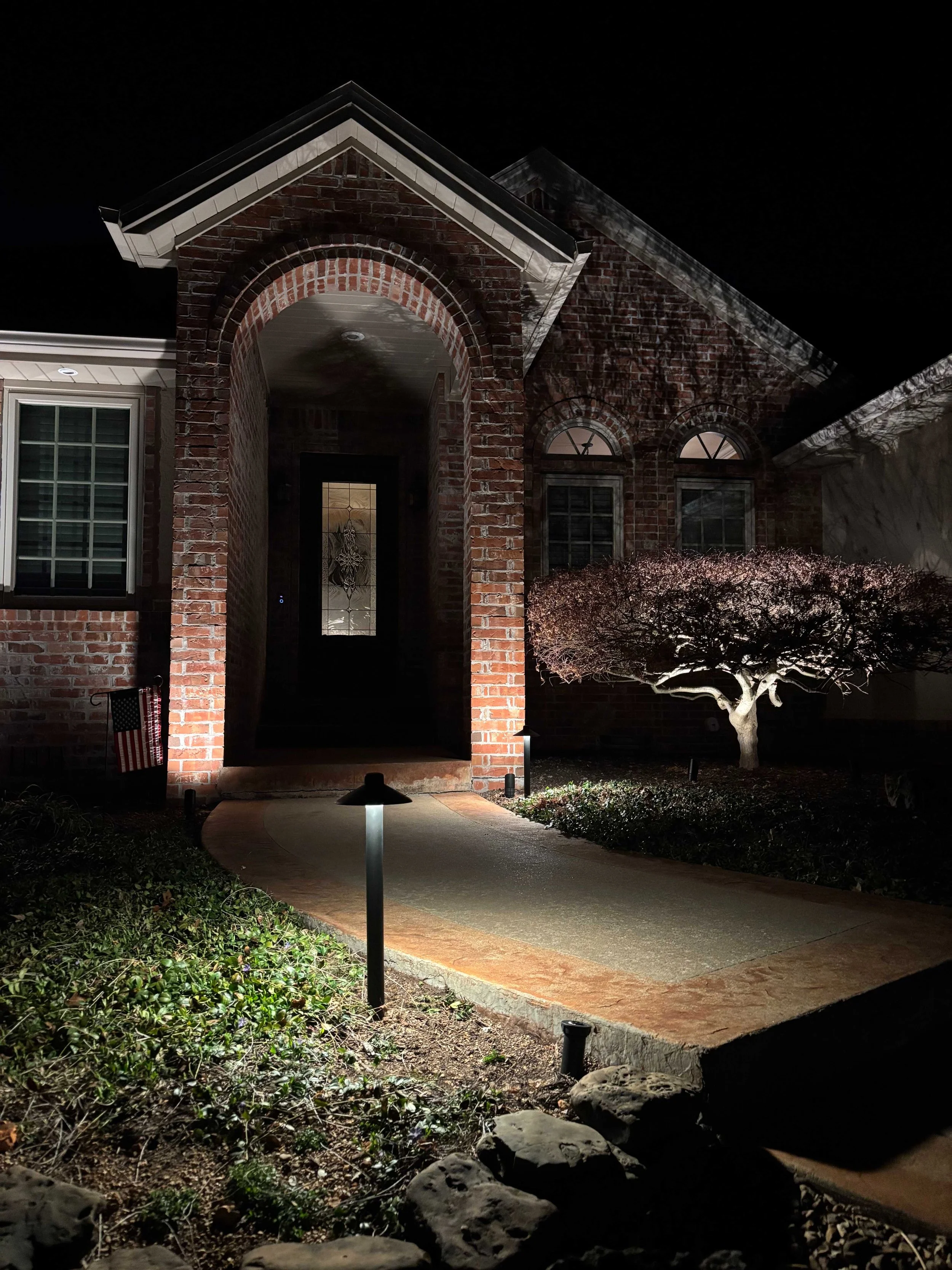 Brick home front entrance at night with professional landscape pathway lighting illuminating the walkway and architectural features in Northwest Arkansas