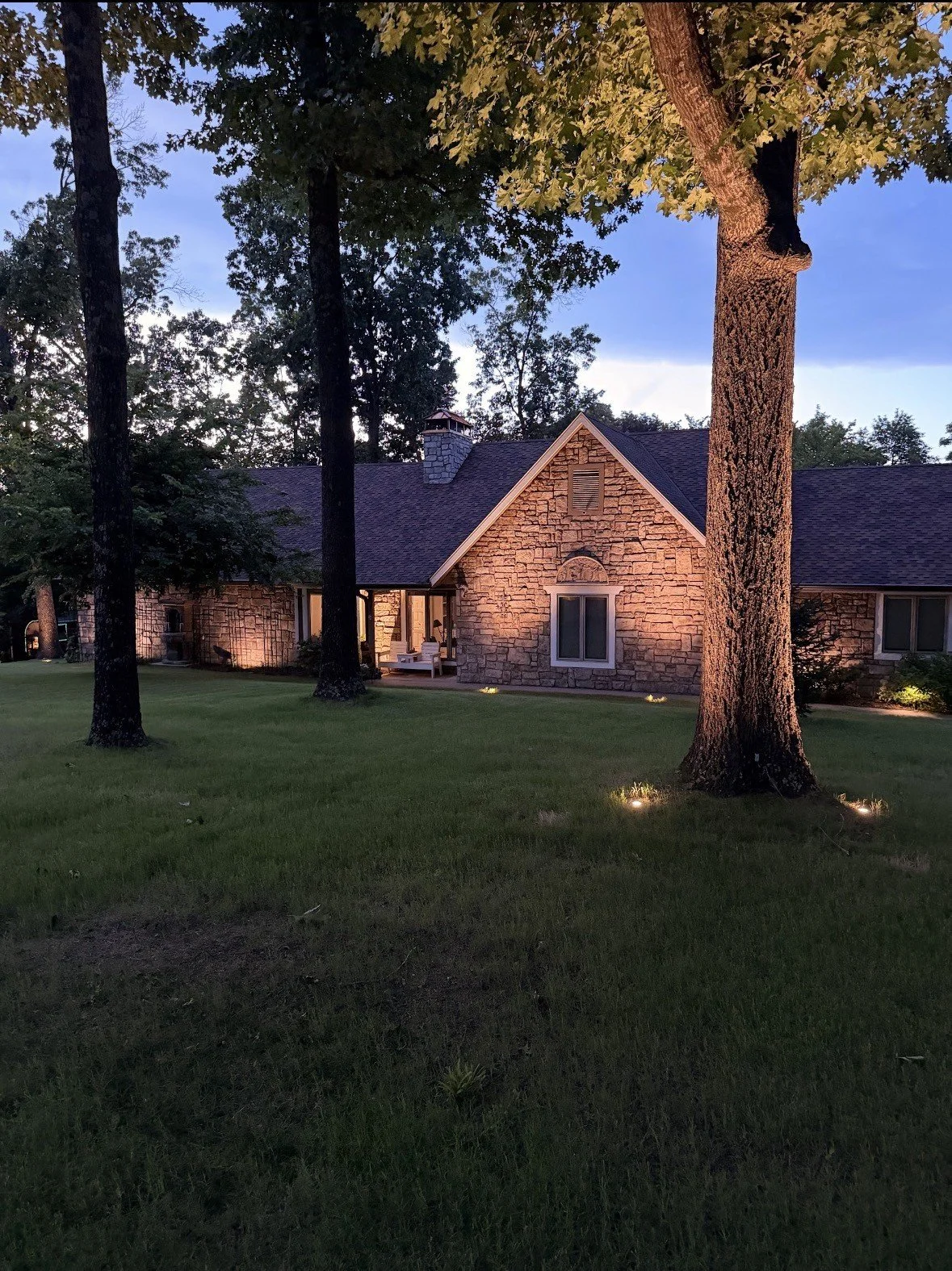 Stone home on Lake Loch Lomond in Bella Vista, AR at dusk with uplit oak trees and a warm facade wash - landscape lighting by Premier Outdoor Lighting and Audio