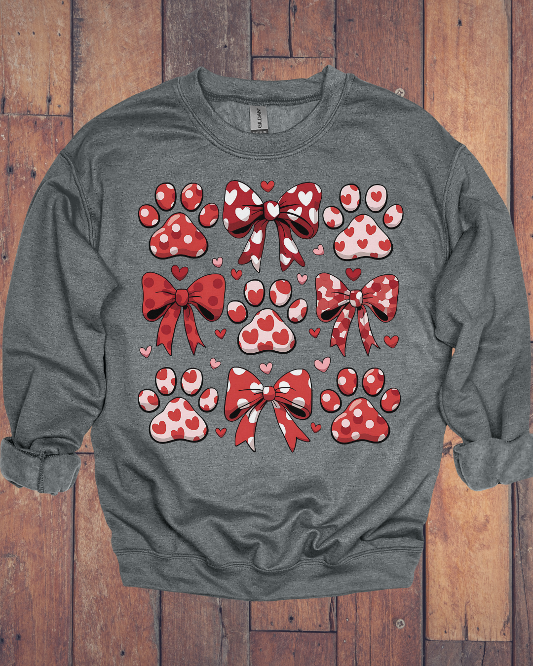Gray sweatshirt with red and pink heart-themed bows, paw prints, and small hearts printed on the front, displayed on a wooden surface.