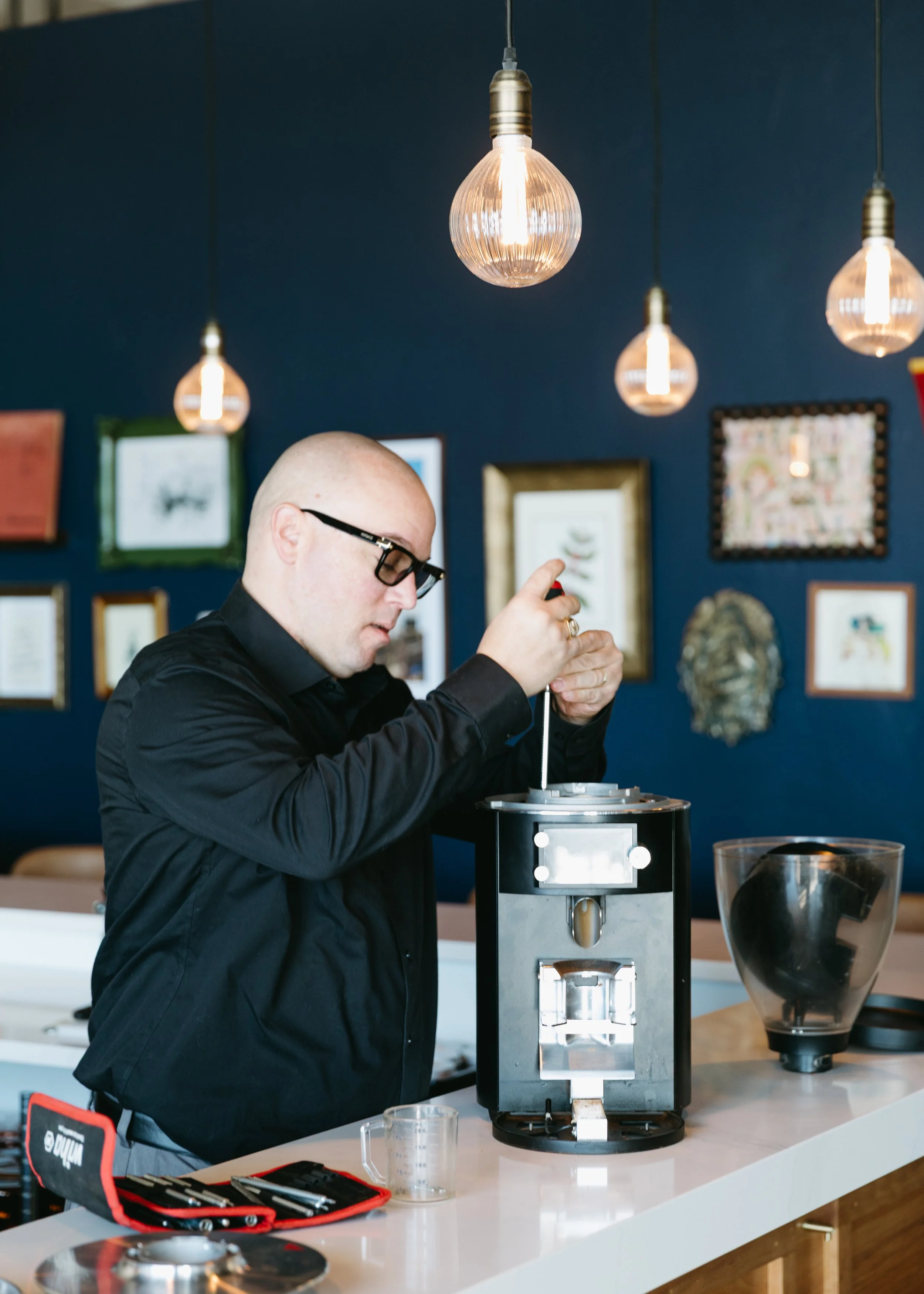 A man with glasses and a black shirt is working on a coffee machine at a counter in a modern, decorated room with framed pictures and hanging lights.